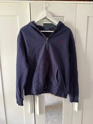  Ralph Lauren tröja - Mörkblå hoodie med dragkedja från Polo Ralph Lauren. Klassisk design med huva, kängurufickor och liten röd logga broderad på bröstet. Tillverkad i mjukt bomullsmaterial och har ribbade muddar vid ärmslut och nederkant.