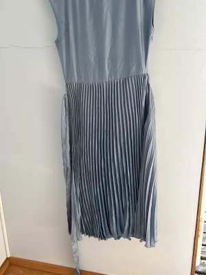Ljusblå plisserad ärmlös klänning - Säljer en elegant ljusblå klänning med plisserad kjol och slät överdel. Klänningen är ärmlös och har en mjuk, rund halsringning. Materialet ser ut att vara lätt och följsamt, perfekt för sommarens tillfällen.