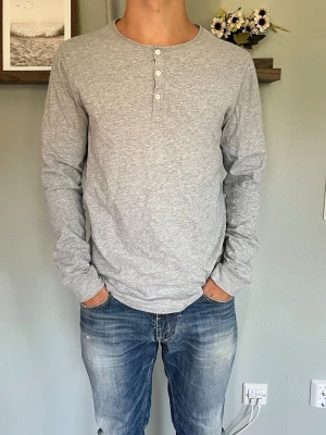 NN07 Henley Långärmad Tröja Grå M - NN07 (No Nationality) henley i gråmelerad bomullsjersey med knappstängning vid halsen och långa ärmar. Skandinaviskt minimalistisk design, mjukt och avslappnat material.  Skick: bra skick, inga hål eller fläckar Storlek: M Modell: 175 cm / 65 kg — sitter true to size på bild