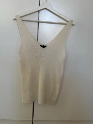 Ribbad beige linnetopp med v-ringning - Snygg ribbad linnetopp i beige med djup v-ringning både fram och bak. Toppen är ärmlös och har en tight passform som framhäver siluetten. Perfekt att styla med jeans eller kjol för en clean och enkel look.