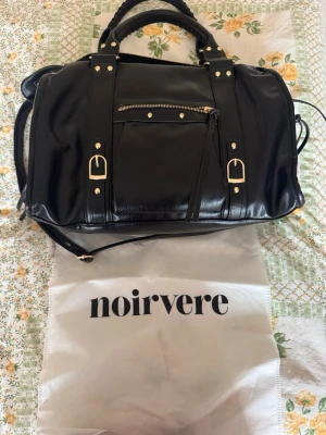 Svart handväska från Noirvere - Stilren svart handväska i glansigt material från Noirvere. Väskan har guldfärgade detaljer, dubbla handtag och en avtagbar axelrem. Framtill finns en ficka med dragkedja och dekorativa spännen. Perfekt för dig som gillar en klassisk men trendig look. Nypris: 1500kr, veganskt läder! Dator får plats!
