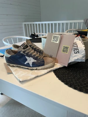 Blå sneakers med vit stjärna - Säljer ett par blå sneakers från Golden Goose med vit stjärna på sidan och grå detaljer. Skorna har texten 'LET A DREAM FLY' på sulan och grå snörning. Ovandelen är i läder och sulan är vit med svart text. Perfekt för dig som gillar unik och cool design.