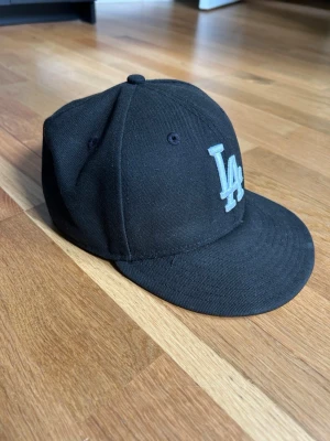 Svart LA-keps från New Era - Snygg svart keps från New Era med LA-logga broderad i vitt framtill. Klassisk modell med böjd skärm och ventilationshål. Perfekt för dig som gillar streetwear och sportig stil. Materialet är tåligt tyg som håller formen. Orginalpris - 500kr 