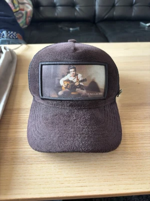 Svart Maggiore truckerkeps med gitarrmotiv - Säljer en svart truckerkeps med mesh baktill och ett stort tygmärke framtill med motiv av en person som spelar gitarr och texten 'MAGGIORE'. Kepsen har böjd skärm och justerbar snapback. Perfekt för dig som gillar musik och unik stil.