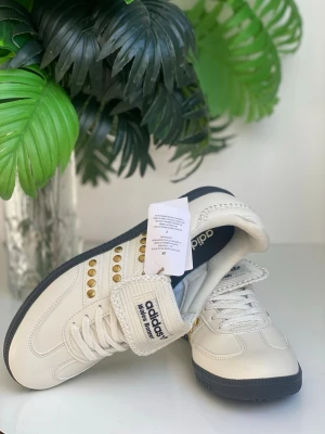 Adidas Wales Bonner vita sneakers - Säljer ett par vita Adidas Wales Bonner sneakers med guldfärgade detaljer och svarta sulor. Skorna har klassisk låg siluett, snörning och kardborreband med tydlig logga. Materialet är slätt läder som ger en clean och stilren look.