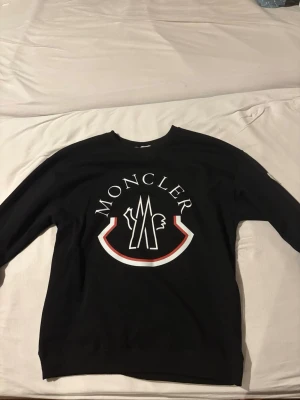 Svart sweatshirt från Moncler - Svart sweatshirt från Moncler med stort vitt och rött logotryck på bröstet och en broderad Moncler-patch på ärmen. Tröjan har rund halsringning och långa ärmar. Perfekt för dig som gillar streetwear och exklusiva märken.