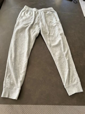 Gråa mjukisbyxor från Nike - Snygga gråa mjukisbyxor från Nike med broderad logga på vänster ben. Byxorna har resår i midjan och muddar vid bensluten för en skön passform. Perfekta för chill dagar eller träning. Tillverkade i mjukt och bekvämt material.