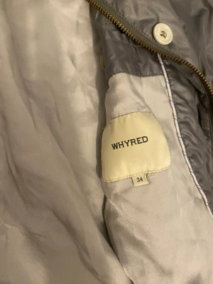 Grå bomberjacka från Whyred - Snygg grå bomberjacka från Whyred med glansigt tyg, ribbade muddar och dragkedja framtill. Jackan har rundad krage och vita knappar längs öppningen. Perfekt för dig som vill ha en stilren och modern look.