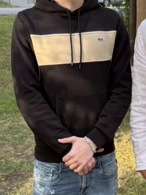 Svart hoodie Lacoste - Snygg svart hoodie från Lacoste med beige panel över bröstet och klassiska krokodilloggan. Huva med dragsko och stor magficka framtill. Perfekt till jeans för en chill och stilren look.