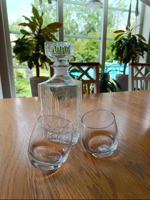 Karaff i glas med två glas - Säljer en stilren karaff i klart glas med fyrkantig form och dekorativ slipning. Till karaffen hör två matchande glas med rundad botten. Perfekt för att servera drycker på ett snyggt sätt. Karaffen har ett rejält lock i samma glasdesign.