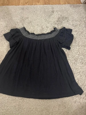 Svart blus från American Eagle - Grå svart  blus från American Eagle med broderad detalj runt halsen och korta volangärmar. Blusen har en lös passform och är tillverkad i mjuk bomull, perfekt för en avslappnad och trendig look.