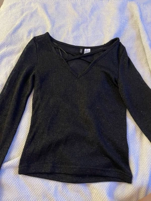 Svart glittrig topp från Divided - Snygg svart glittrig topp från Divided i storlek XS. Toppen har lång ärm, v-ringning med dekorativa band över bröstet och en slim passform. Perfekt för dig som vill ha något extra till outfiten!