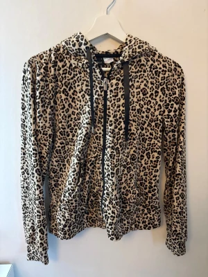 Leopardmönstrad hoodie med dragkedja - Säljer en leopardmönstrad hoodie med dragkedja framtill och svarta snören i huvan. Hoodien har långa ärmar och är gjord i ett mjukt, stretchigt material. 