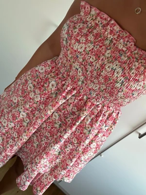 Axelbandslös blommig kortklänning - Söt axelbandslös kortklänning med rosa och vita blommor. Klänningen har smock upptill och ett lätt utställt kjolparti. Perfekt för varma dagar och ger en romantisk vibe.