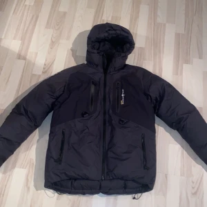 Svart dunjacka från Sail Racing - Säljer en svart dunjacka från Sail Racing med GORE-TEX-material. Jackan har en justerbar huva och flera praktiska dragkedjefickor. Perfekt för kalla äventyr med sin robusta design och varma fyllning. Den har en snygg patch med 'Antarctica Expedition' på ärmen. Nypris ca 9000kr