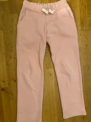 Ljusrosa mjukisbyxor från russemerch - Mysiga ljusrosa mjukisbyxor med resår i midjan och en söt rosett framtill. Byxorna har två fickor och är gjorda i ett mjukt, fleeceliknande material som är perfekt för chill dagar. Passar dig som gillar en relaxed och comfy stil.