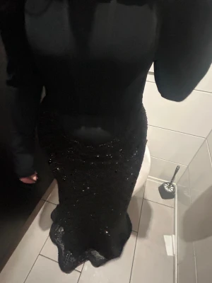 Svart offshoulder långklänning - Elegant svart långklänning med offshoulder-design och långa ärmar. Kjolen är glittrig och figurnära, vilket ger en lyxig känsla. Perfekt för dig som vill sticka ut med en stilren och glamorös look.