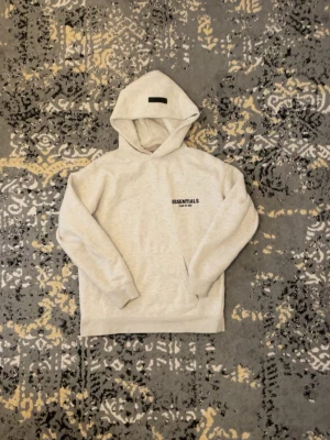 Essentials Fear of God hoodie beige - Snygg beige hoodie från Essentials Fear of God med svart logga på bröstet och stor text på ryggen. Hoodien har huva och känguruficka framtill. Perfekt för dig som gillar streetwear och vill ha en clean look.