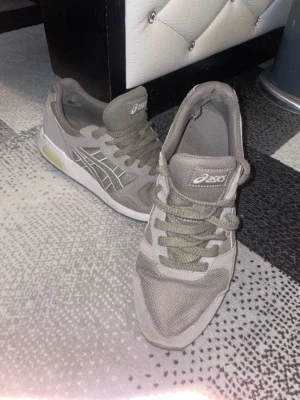 Gråa Asics sneakers med mesh och mocka - Säljer ett par gråa Asics sneakers med klassisk logga på sidan. Skorna har ovandel i mesh och mocka, snörning och vit sula med beige detalj vid hälen. Perfekta för dig som gillar sportig och avslappnad stil.