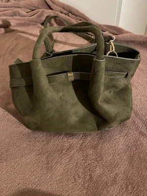 Olivgrön handväska i mockaimitation - Snygg olivgrön handväska i mjuk mockaimitation med både handtag och axelrem. Väskan har en dekorativ rem med metallspänne framtill och rymligt huvudfack. Perfekt för dig som vill ha en stilren och trendig accessoar.