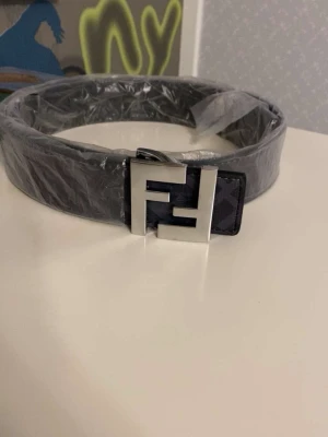 Svart fendi bälte med silverfärgat spänne - Snyggt svart fendi bälte med ett stort, fyrkantigt spänne i silverfärg. Bältet har en stilren design och passar perfekt till jeans eller kostymbyxor. Spännet har en unik geometrisk form som ger bältet en modern touch.