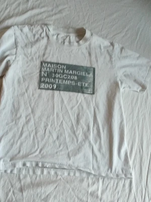 Maison Martin Margiela vit t-shirt - Vit t-shirt med tryck på bröstet från Maison Martin Margiela. Trycket är i grått och vitt och har texten 'PRINTEMPS-ETE 2009'. T-shirten har rund hals och korta ärmar, tillverkad i mjukt bomullstyg.