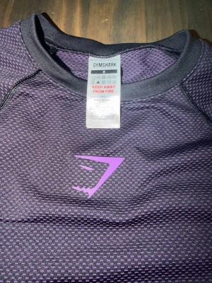 Onyx • Gymshark • lila • Small - Snygg lila och svart träningströja från Gymshark med korta ärmar och rund hals. Tröjan har ett mönstrat meshparti upptill och Gymshark-logga i lila på bröstet. Tillverkad i ett stretchigt funktionsmaterial som andas, perfekt för gymmet eller löpning.