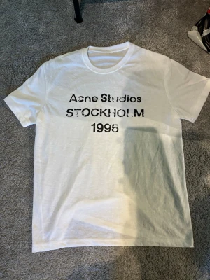 Vit Acne Studios t-shirt Stockholm - Vit t-shirt från Acne Studios med svart tryck framtill där det står 'Acne Studios STOCKHOLM 1996'. Klassisk rund hals och korta ärmar. Tillverkad i mjuk bomull för en skön och avslappnad look.