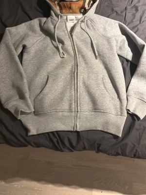 Grå hoodie med kamouflagemönstrad huva - Säljer en grå hoodie med dragkedja och fickor framtill. Huvan har ett unikt kamouflagemönstrat foder och justerbara snören. Tröjan har ribbade muddar vid ärmar och nederkant. Perfekt för dig som gillar enkel men snygg streetwear.