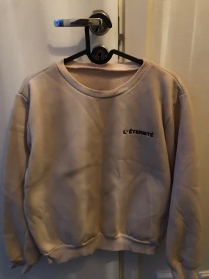 Beige sweatshirt med tryck - Beige sweatshirt med rund halsringning och trycket 'L'ÉTERNITÉ' på bröstet. Tröjan har ribbade muddar vid ärmslut och nederkant, samt en avslappnad passform. Perfekt för en chill och stilren look.