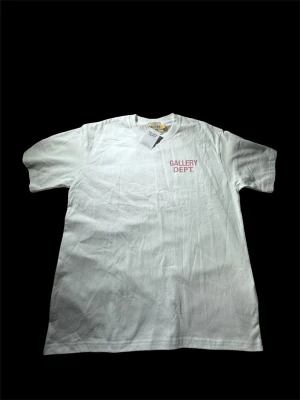 Gallery Dept. T-shirt – Vit/Rosa – Storlek M  - Säljer en stilren t-shirt från Gallery Dept. i storlek M. Den har en klassisk vit design med den ikoniska Gallery Dept.-loggan i rosa på bröstet, vilket ger en clean och modern look som passar till i princip allt. T-shirten är tillverkad i mjuk bomull med en bekväm passform och passar perfekt till både jeans, shorts eller cargos. En snygg streetwear-klassiker som funkar året om. Skicka gärna ett meddelande om du har frågor eller vill ha fler bilder - Svarar snabbt!