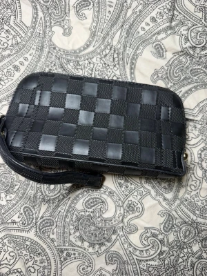 Svart plånbok med rutmönster - Snygg svart plånbok med ett schackrutigt mönster i både matt och blank finish. Plånboken har handledsrem och dragkedja runt om. Tillverkad i konstläder med stilren design som passar perfekt för att hålla koll på kort och kontanter.