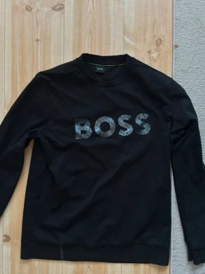 Svart sweatshirt från BOSS - Svart sweatshirt från BOSS med stort logotryck i grått och svart mönster över bröstet. Klassisk rund halsringning och långa ärmar. Perfekt för en stilren och avslappnad look.