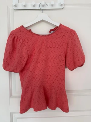 Korallfärgad blus med puffärm - Söt korallfärgad blus med korta puffärmar och volang nedtill. Blusen har ett diskret hålmönster och rund halsringning. Stängs med knapp i nacken. Perfekt för dig som gillar romantiska detaljer och en feminin look.💕