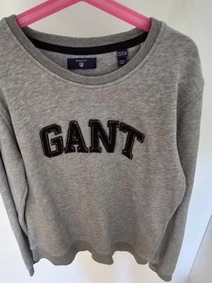 Grå sweatshirt från GANT 158/164 - Klassisk grå sweatshirt från GANT med stor svart logotyp i college-stil framtill. Tröjan har rund halsringning och är tillverkad i mjuk bomullsmix som passar perfekt till vardagslooken.