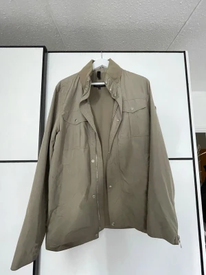 Oliv jacka från J.Lindeberg - Stilren beige jacka från J.Lindeberg med klassisk krage, tryckknappar och två bröstfickor med lock. Jackan har lång ärm och är en regn jacka. Perfekt för dig som gillar enkel och stilren look.
