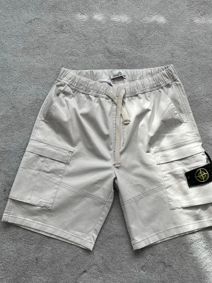Beiga cargoshorts från Stone Island - Säljer ett par beiga cargoshorts från Stone Island med klassisk patch på benet. Shortsen har flera fickor, dragsko i midjan och är tillverkade i mjuk bomull. Perfekta för sommaren och riktigt snygga till streetwear-stilen.