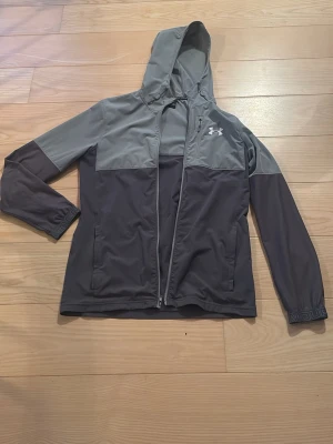 Grå vindjacka från Under Armour - Snygg tvåfärgad vindjacka från Under Armour i ljus- och mörkgrått. Jackans dragkedja är lite sönder men den går att stänga, man kan inte öppna dragkedjan längst ner ibland, men annars är skicket perfekt 