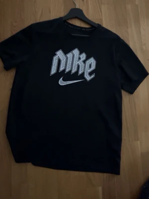 Svart Nike t-shirt med tryck - Säljer en svart t-shirt från Nike med stort grafiskt tryck i vitt och grått på bröstet. Klassisk rund hals och korta ärmar. Perfekt för en sportig och avslappnad stil.