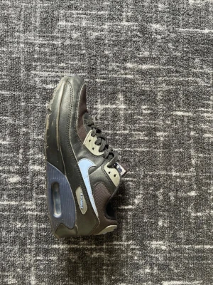 Nike Air Max 90 svart/blå sneakers - Säljer ett par Nike Air Max 90 sneakers i svart och blått med klassisk synlig luftbubbla i sulan. Skorna har snörning och är tillverkade i en mix av syntet och textil. Swoosh-logga på sidan och detaljer i grått och blått. Perfekta för dig som gillar streetwear.