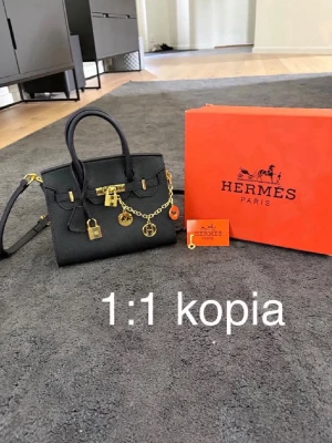 Svart Birkin handväska med gulddetaljer - Snygg svart handväska i klassisk modell med guldfärgade metalldetaljer, hänglås och kedja. Väskan har både handtag och axelrem samt dekorativa berlocker. Perfekt för dig som gillar lyxig och trendig stil.
