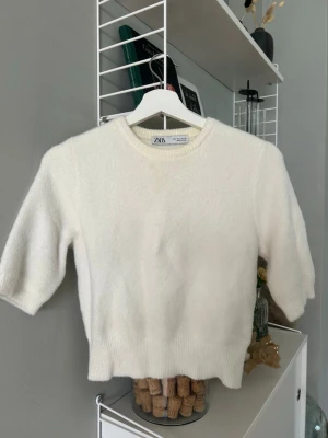 Vit stickad tröja från Zara - Mysig vit stickad tröja från Zara med rund halsringning och halvlånga ärmar. Tröjan har en enkel och stilren design med ribbade muddar vid ärmslut och nederkant. Perfekt för en clean look.