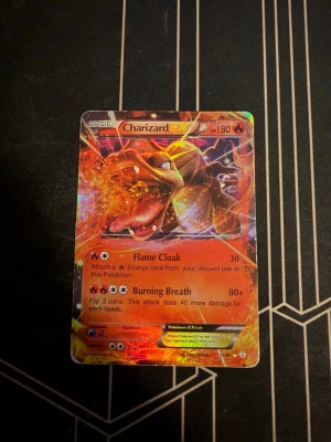 Charizard EX Pokémonkort - Säljer ett holografiskt Charizard EX Pokémonkort med 180 HP. Kortet har en eldig design i rött, orange och gult med attacker som Flame Cloak och Burning Breath. Perfekt för samlare eller Pokémonfans som vill utöka sin kortsamling.