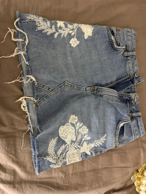 Blå jeansshorts med broderade blommor - Säljer ett par blå jeansshorts med råa kanter och vita broderade blommor framtill och baktill. Shortsen har klassisk femficksdesign, bälteshällor och dragkedja med knapp. Perfekta för sommaren och ger en cool, avslappnad vibe.