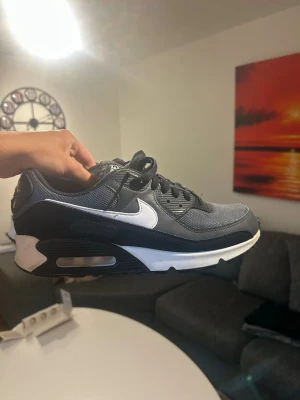 Nike Air Max 90 svart/vit sneaker - Snygga Nike Air Max 90 sneakers i svart och vitt med klassisk siluett. Ovandelen är i mesh och läder, med vit swoosh på sidan och synlig Air-enhet i sulan. Skorna har svarta skosnören och en robust yttersula för bra grepp.