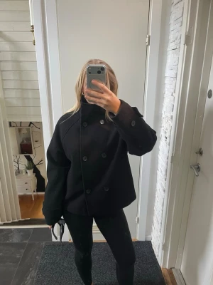 Svart dubbelknäppt rock - Säljer en svart, kortare rock med dubbelknäppning och hög krage. Rocken har en oversized passform och vida ärmar, vilket ger en modern och avslappnad look. Perfekt för dig som gillar stilrena och minimalistiska plagg. Original pris 999 kr