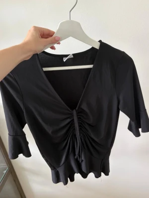 Svart topp från Gina Tricot - Svart topp från Gina Tricot med v-ringning och snörning framtill. Toppen har trekvartsärm med volangkant och är croppad i modellen. 