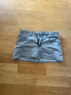 Grå jeanskjol med fickor - Snygg grå jeanskjol i kort modell med klassiska fem fickor, bälteshällor och knappstängning fram. Kjolen har en rak passform och är tillverkad i denim med en tvättad look. Perfekt för dig som gillar en enkel men trendig stil.