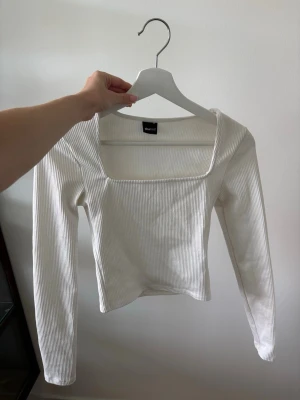 Vit ribbad långärmad topp - Trendig vit ribbad topp med lång ärm och fyrkantig urringning. Toppen har en croppad passform och är tillverkad i ett mjukt och stretchigt material.