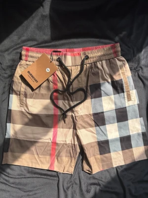 Badbyxor Burberry - Säljer ett par klassiska badbyxor från Burberry i beige, svart, vitt och rött med det ikoniska rutmönstret. De har elastisk midja med svart dragsko och sidofickor. Perfekta för stranden eller poolen och ger en lyxig känsla till din sommarstil. Storlek M